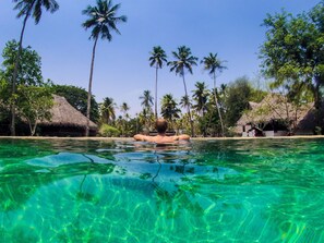 Une piscine aux eaux cristallines où flotte une personne, entourée de palmiers et de petites huttes aux toits de chaume.