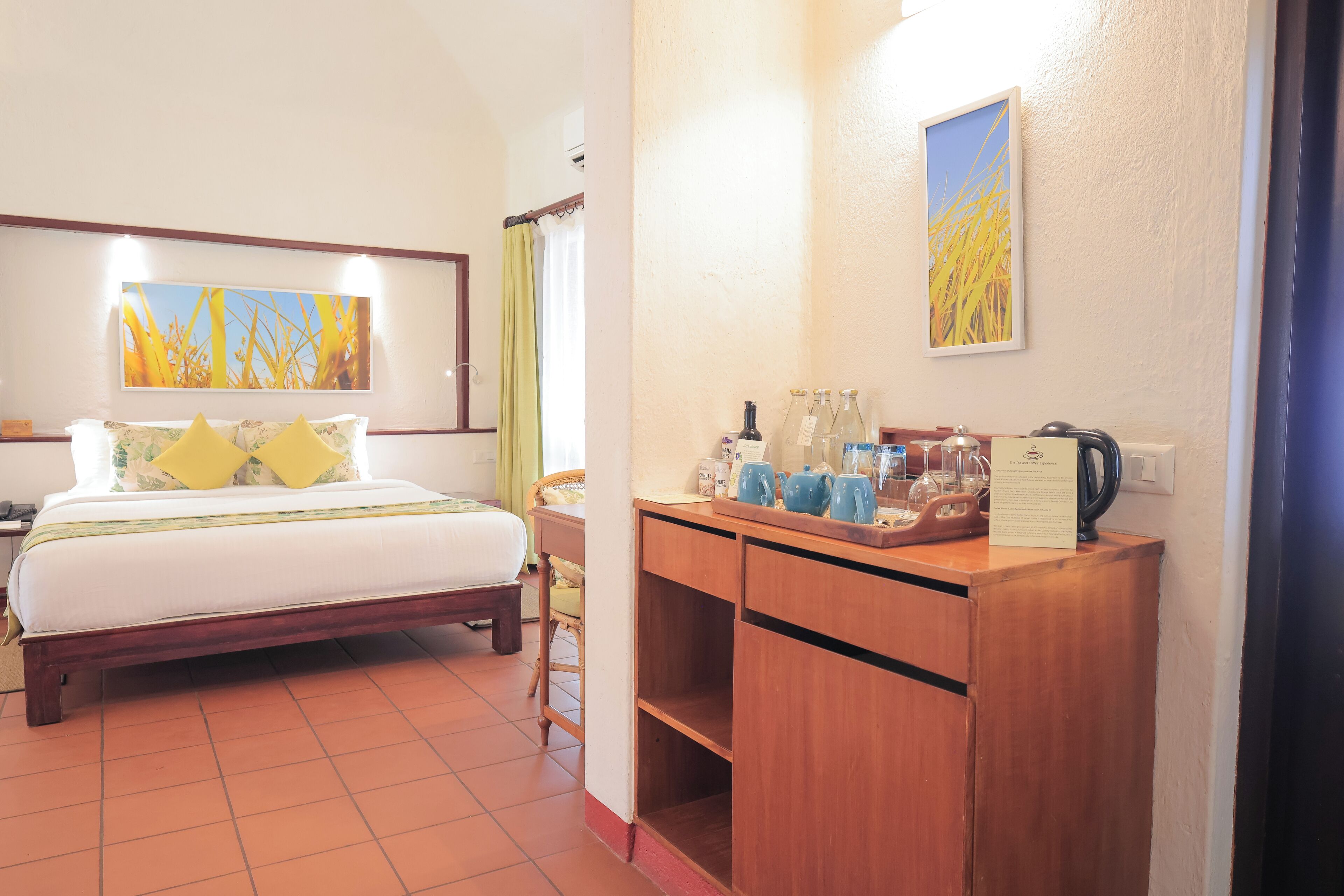 Habitación de hotel con una cama, un escritorio de madera con tetera, teteras y vasos, y dos cuadros en la pared.