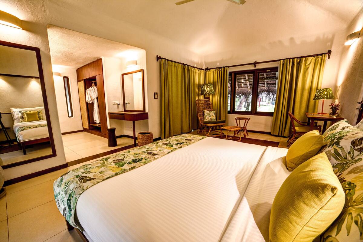 A hotel room with a large bed, a sitting area with a chair and table, a mirror, and a view of the outdoors through a window.
