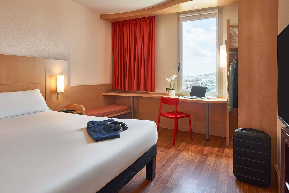 Habitación de hotel con cama, escritorio con computadora portátil, silla, maleta y ventana con vista a la ciudad.