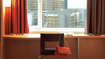 Una habitación de hotel con vistas a edificios, una silla y un bolso rojo en el alféizar de la ventana.