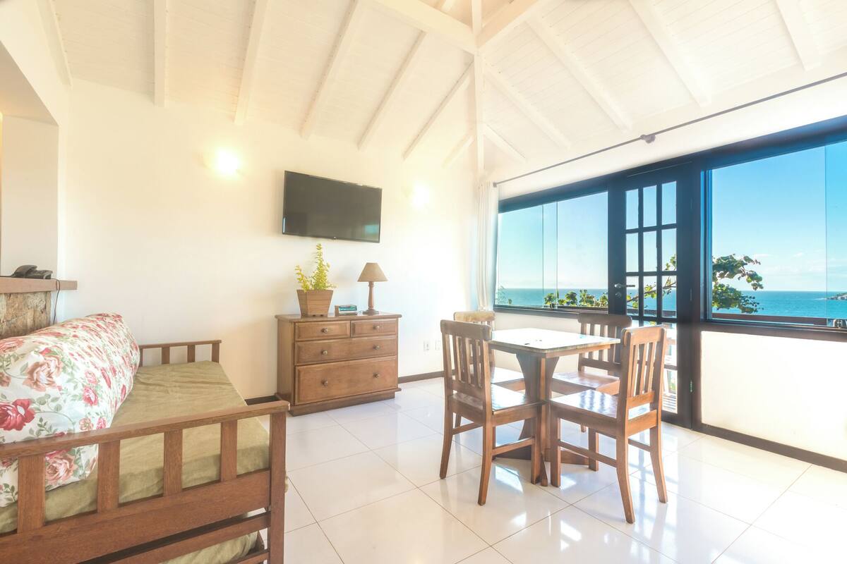 Ein Zimmer mit einem Bett, einem Holzschrank, einem Tisch mit Stühlen, einem an der Wand befestigten Fernseher und einem großen Fenster mit Meerblick.