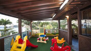 Children can play on a slide, rocking horse, and table in this outdoor play area with a view of palm trees and a body of water.