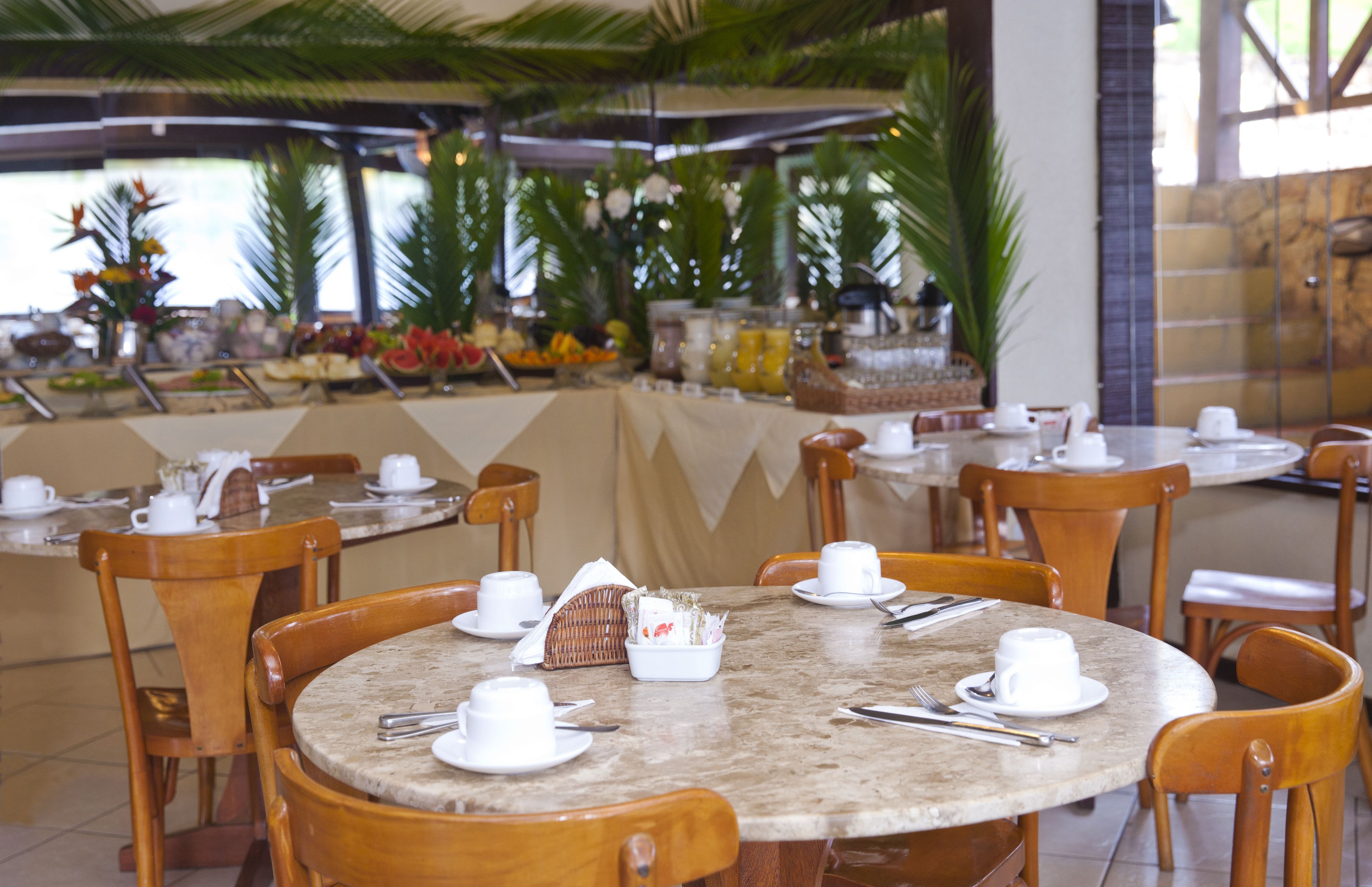A restaurant with round tables set for dining, wooden chairs, and a buffet area with various food items.