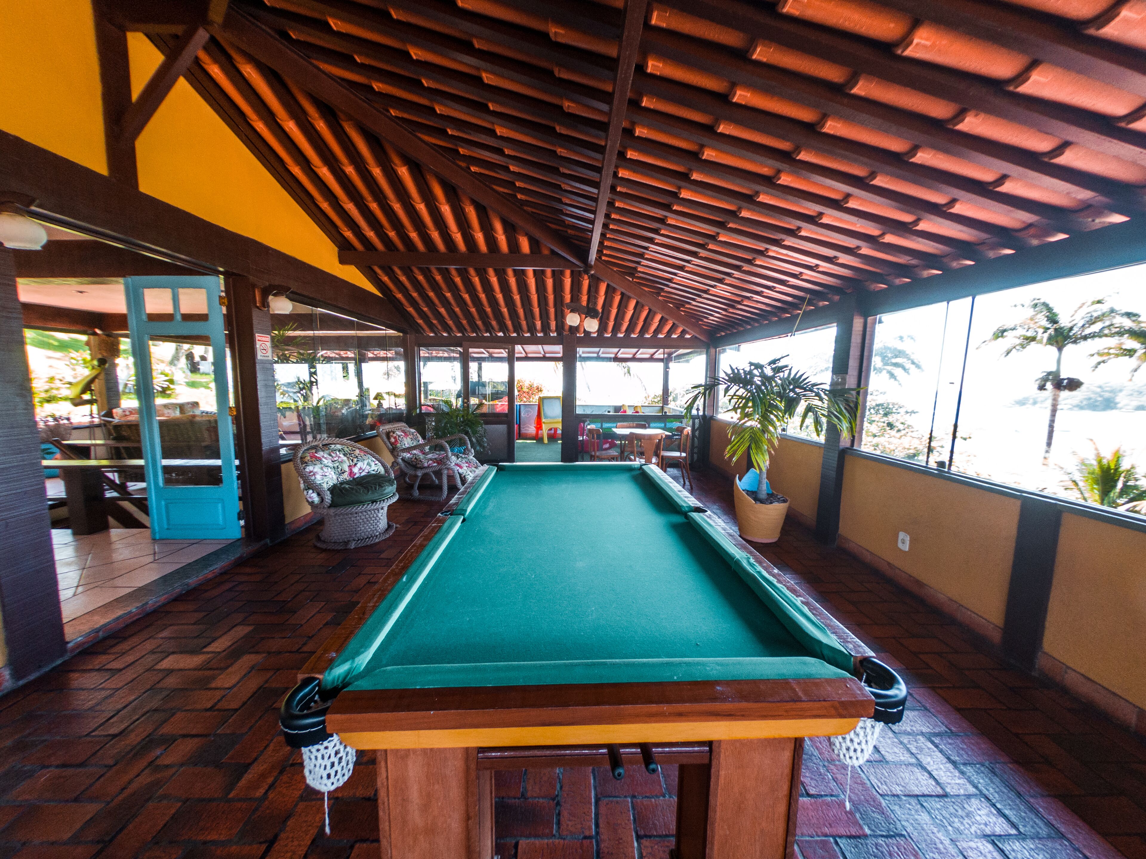 A pool table in a covered area with chairs and tables, large windows, and a view of palm trees.