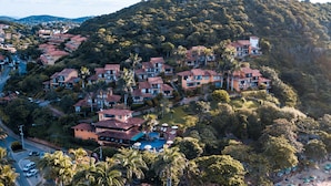 Vista aérea de un complejo turístico costero con varias edificaciones, una playa con sombrillas y tumbonas, y una piscina.