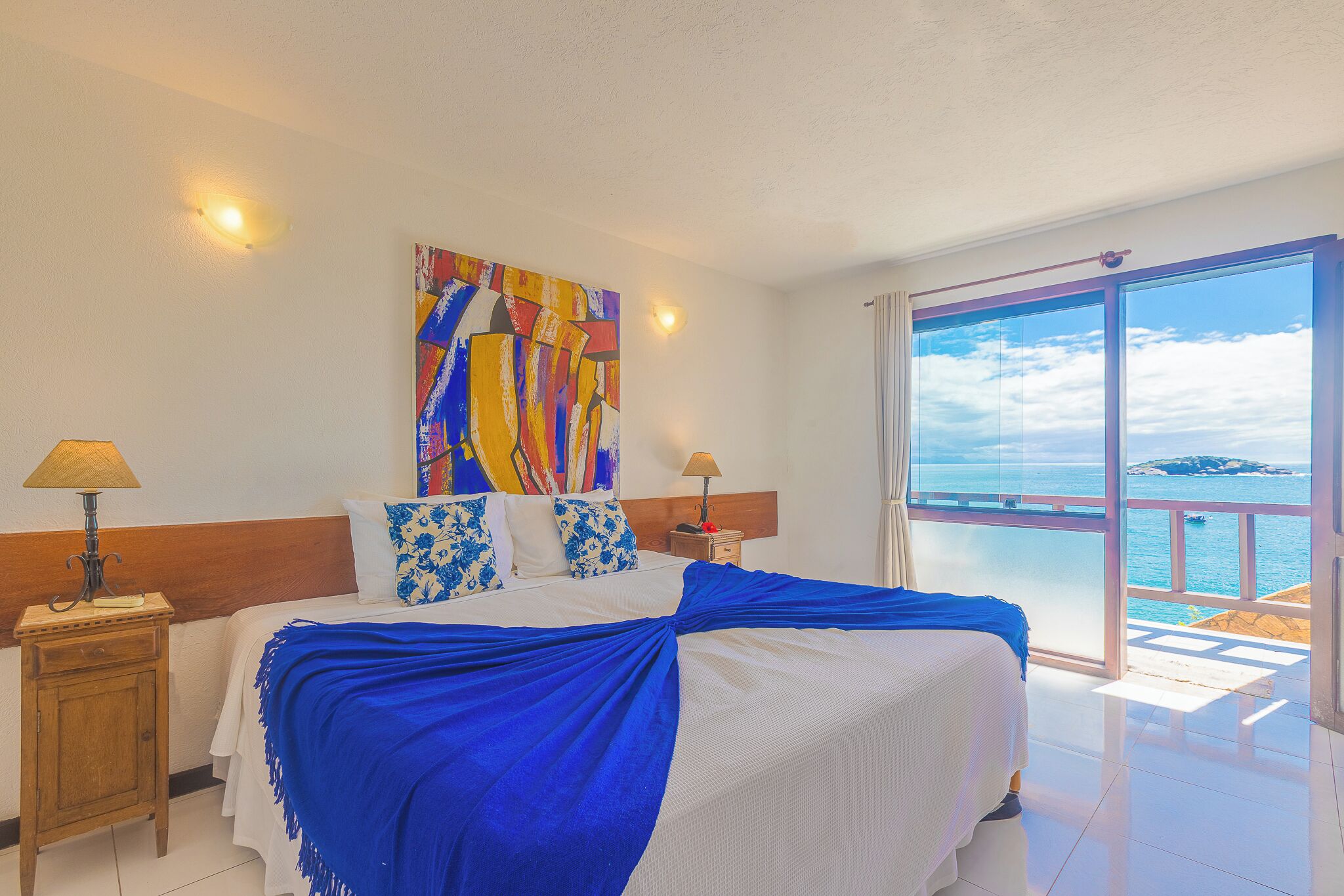 A hotel room with a bed, a painting, a nightstand, a lamp, and a large window with a view of the sea.