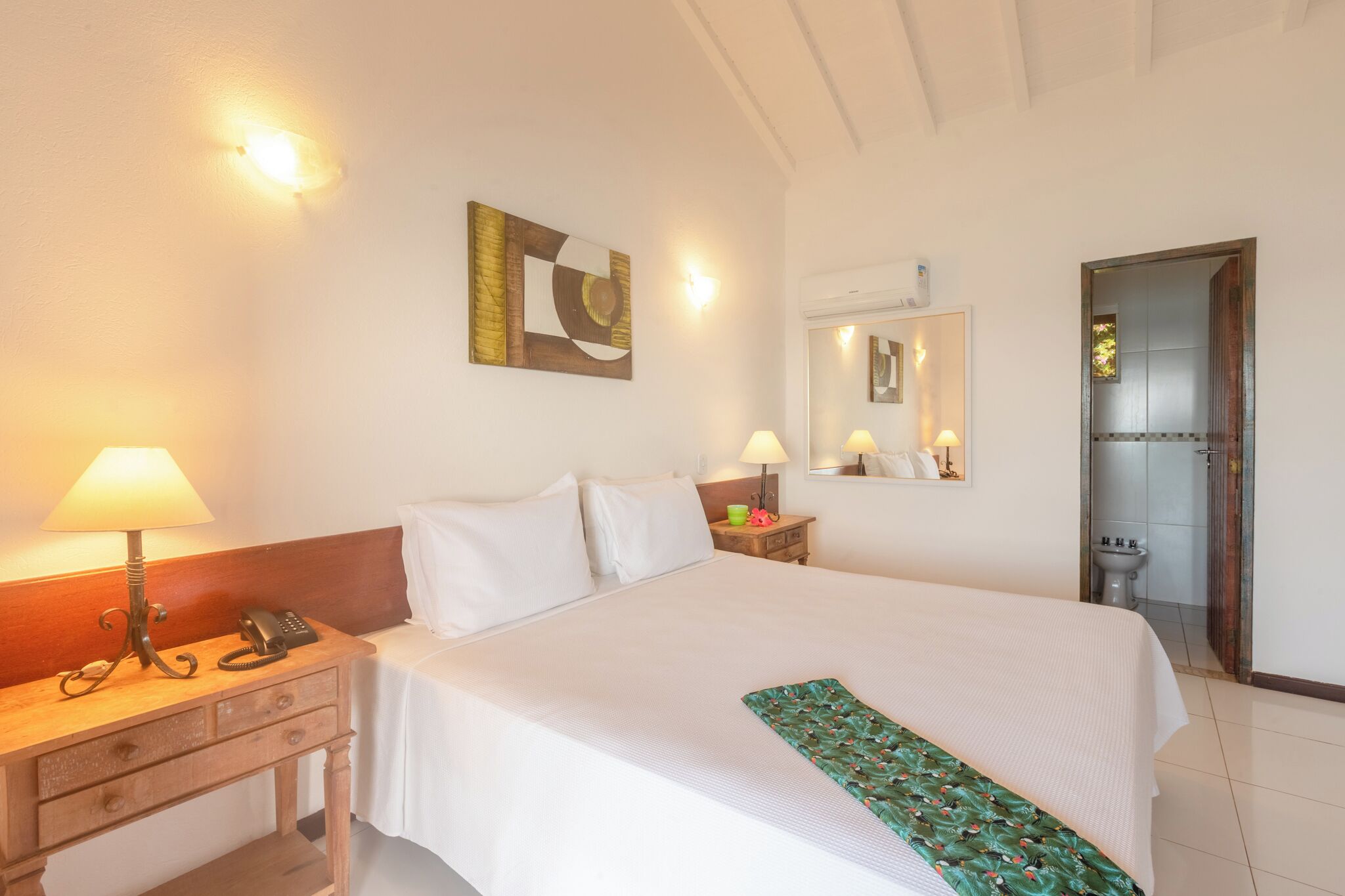 Habitación de hotel con cama, lámparas de noche, teléfono, mesita de noche, espejo y puerta que da a un baño.