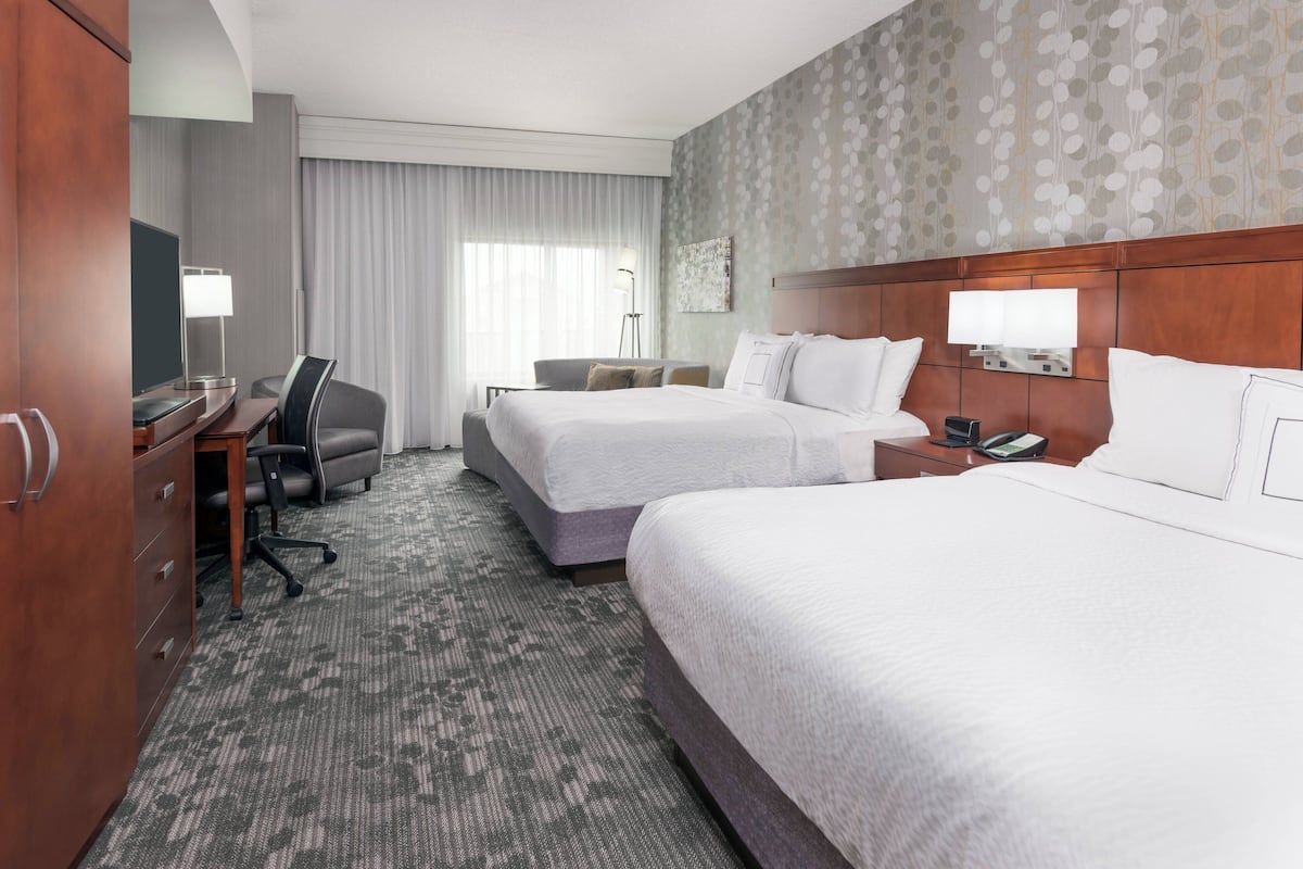 A hotel room with two beds, a desk, a chair, a TV, and a lamp.
