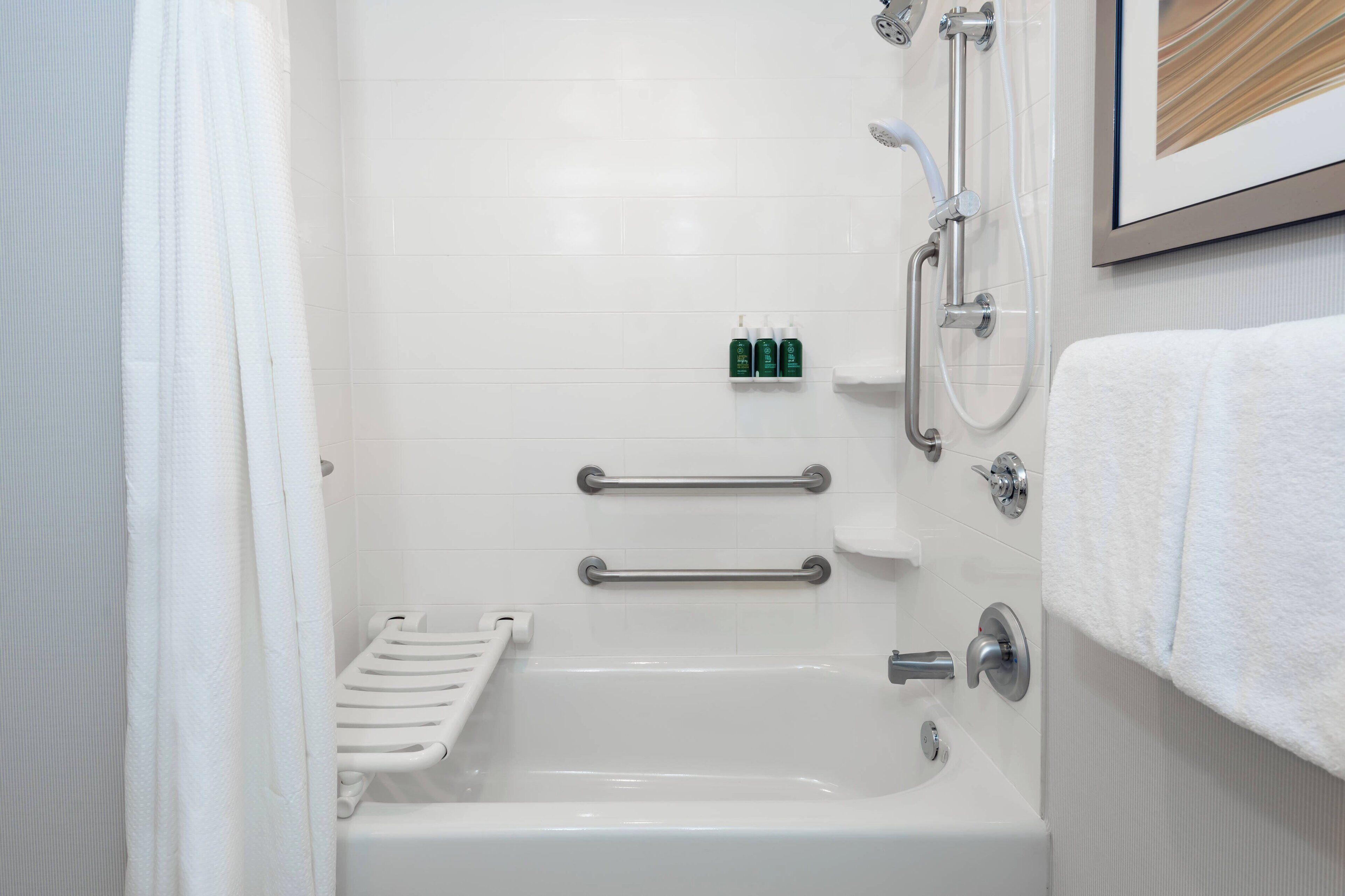 Un baño moderno con una bañera blanca, barras de apoyo, cabezal de ducha y un cuadro en la pared.