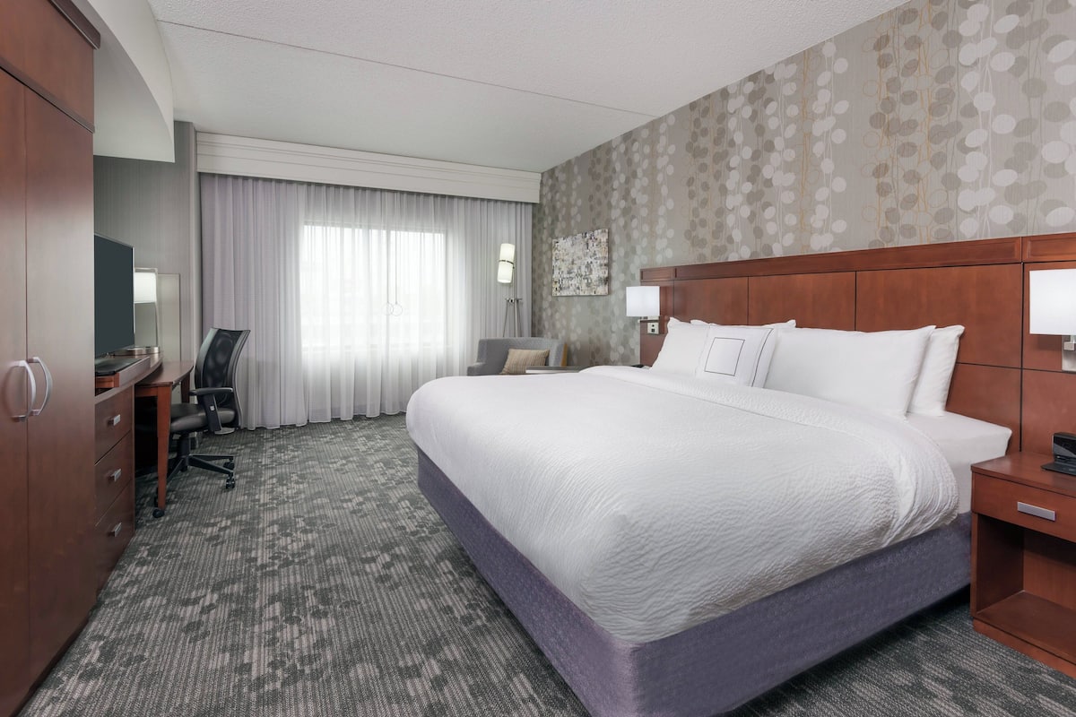 A hotel room with a large bed, a desk with a chair, a wardrobe, and a window with curtains.