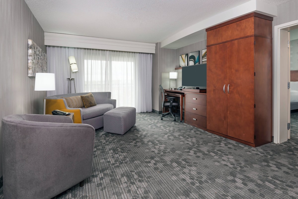 A hotel room with a grey sofa, a small ottoman, a desk with a chair, a wardrobe, and a large window with curtains.