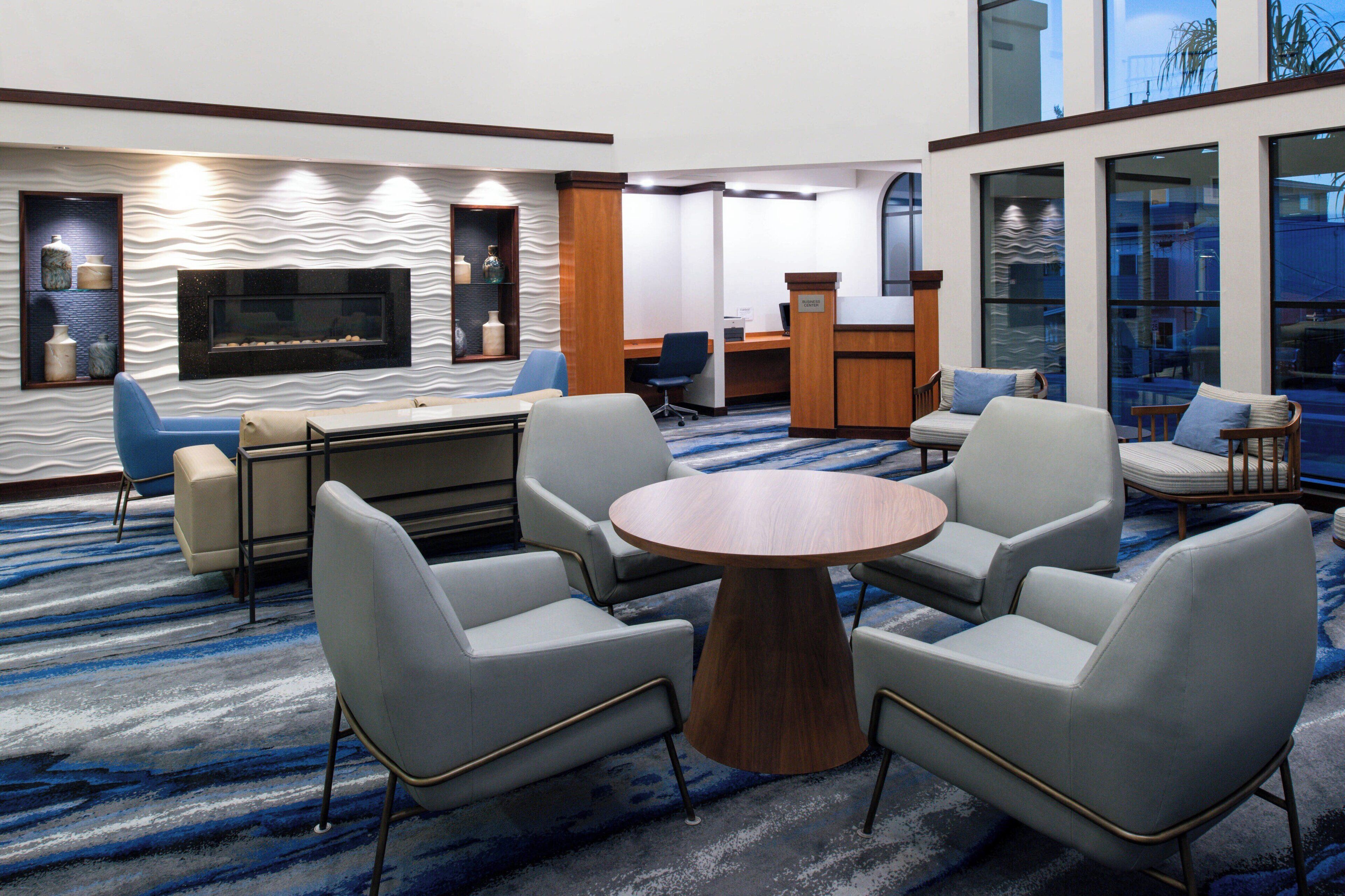 A modern hotel lobby with a round wooden table surrounded by four chairs, a sofa, a fireplace, and a reception desk.