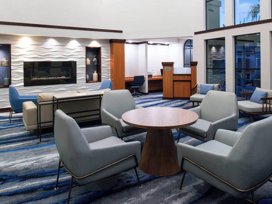 A modern hotel lobby with a round wooden table surrounded by four chairs, a sofa, a fireplace, and a reception desk.