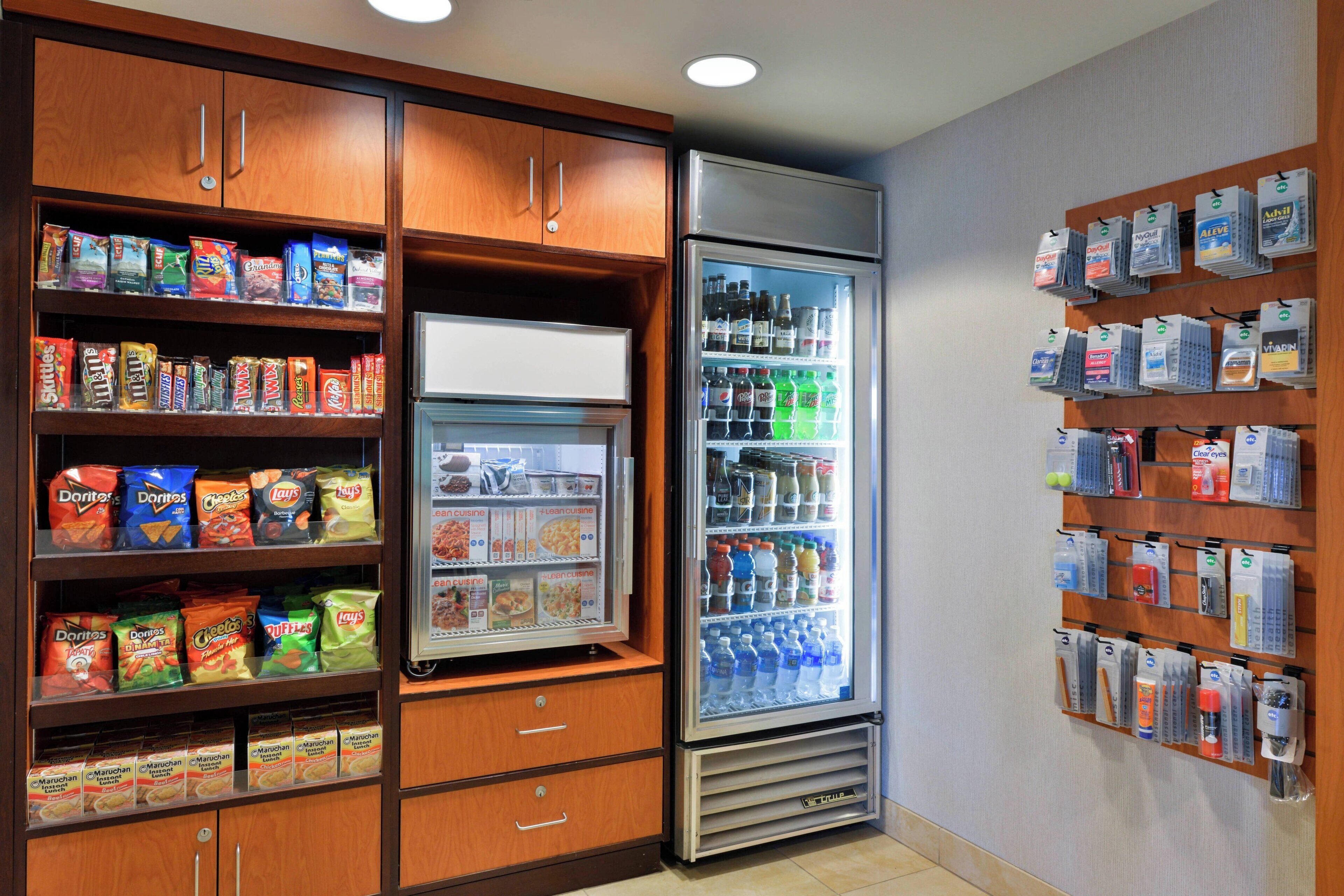 Un refrigerador con aperitivos y bebidas, repleto de diversas opciones, rodeado de armarios de madera y una vitrina empotrada con agua embotellada y otras bebidas.