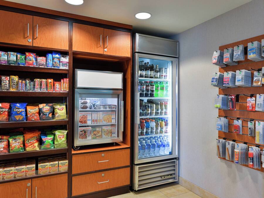 Un refrigerador con aperitivos y bebidas, repleto de diversas opciones, rodeado de armarios de madera y una vitrina empotrada con agua embotellada y otras bebidas.