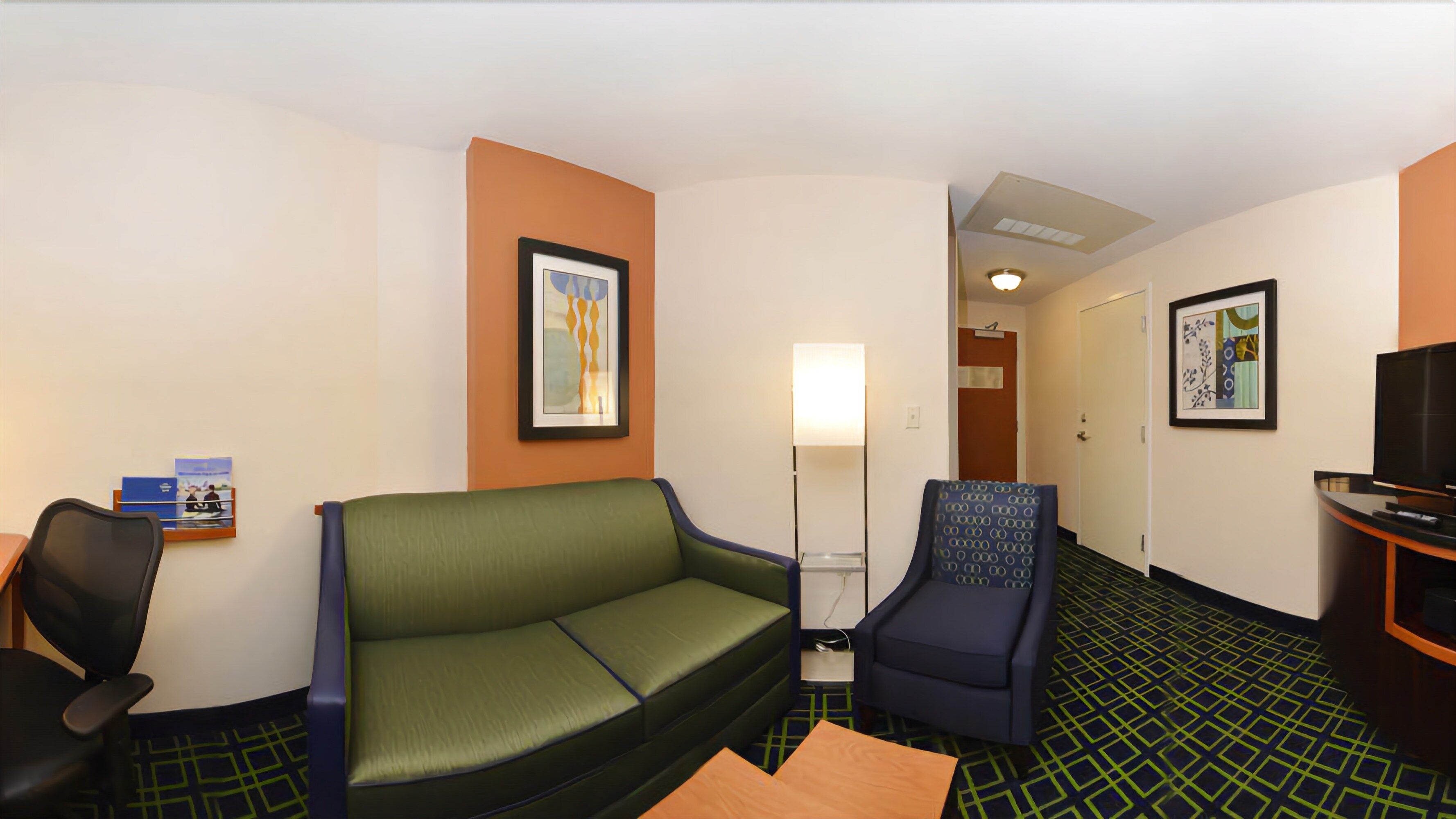 Een hotelkamer met een groene bank, een blauwe fauteuil, een houten tafel, een televisie en lijstwerken aan de muren.