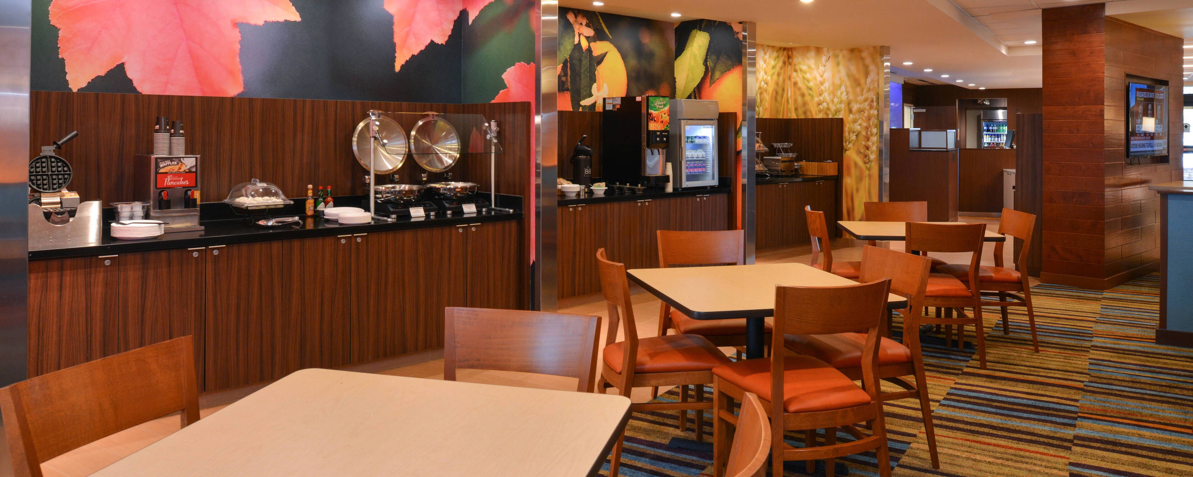 Un comedor de hotel con mesas y sillas de madera, una barra de buffet con diversas opciones de comida, y un mostrador con una máquina de café.