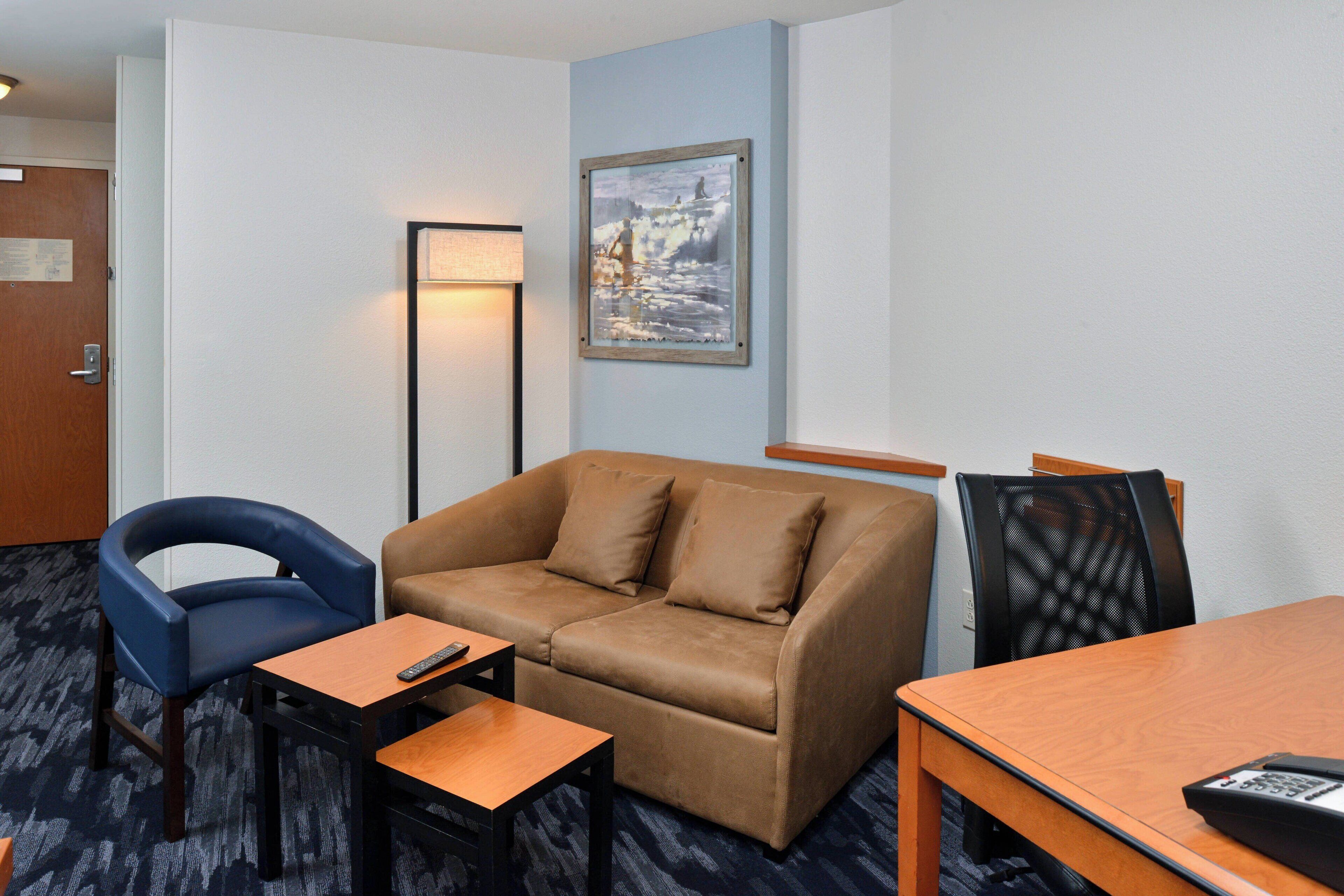 A hotel room with a brown sofa, a blue armchair, a small wooden table, and a desk with a remote control.