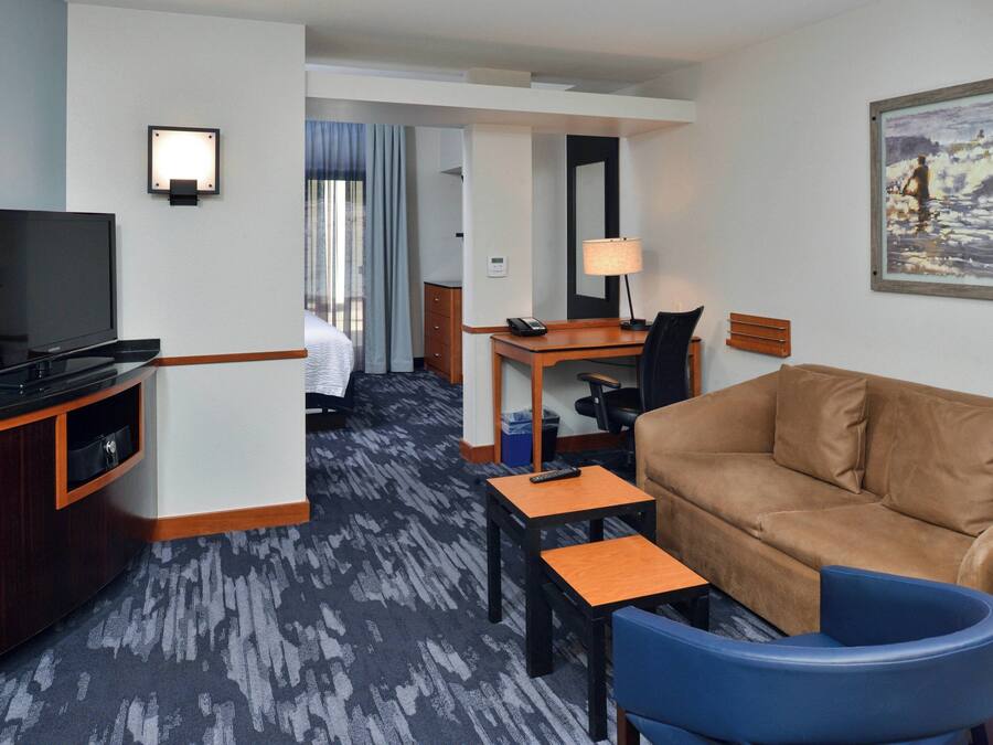 Una habitación de hotel con una televisión de pantalla plana, un sofá de cuero marrón, un sillón azul, una pequeña mesa de madera y un escritorio con una lámpara.