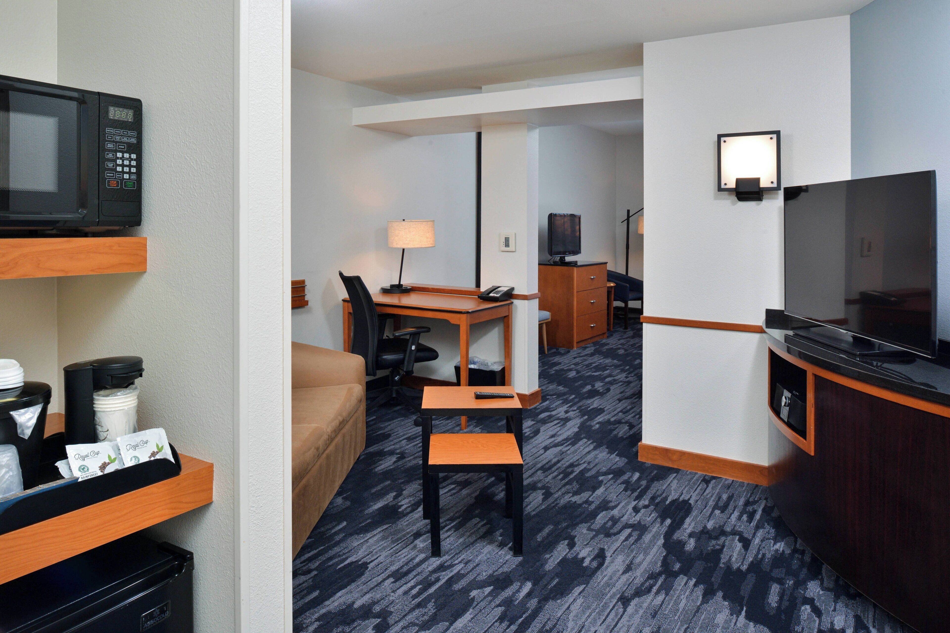 A hotel room with a kitchenette, a TV, a desk with a chair, a lamp, and a bed.