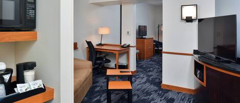 A hotel room with a kitchenette, a TV, a desk with a chair, a lamp, and a bed.