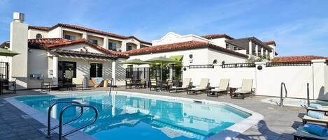 A swimming pool with lounge chairs and umbrellas, surrounded by a building with a tiled roof and white walls.