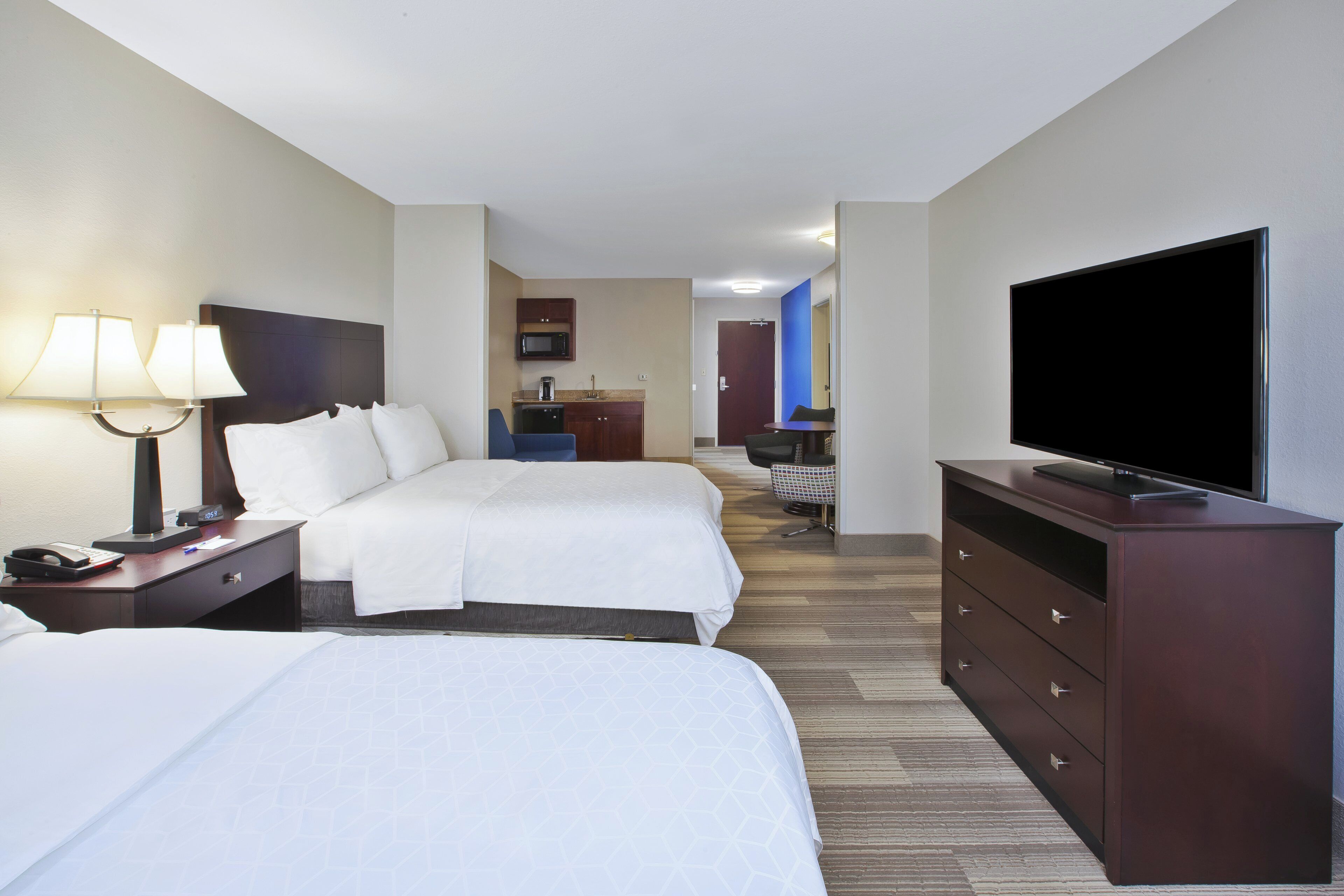 Habitación de hotel con dos camas, televisión, escritorio con lámpara, cómoda y una kitchenette al fondo.
