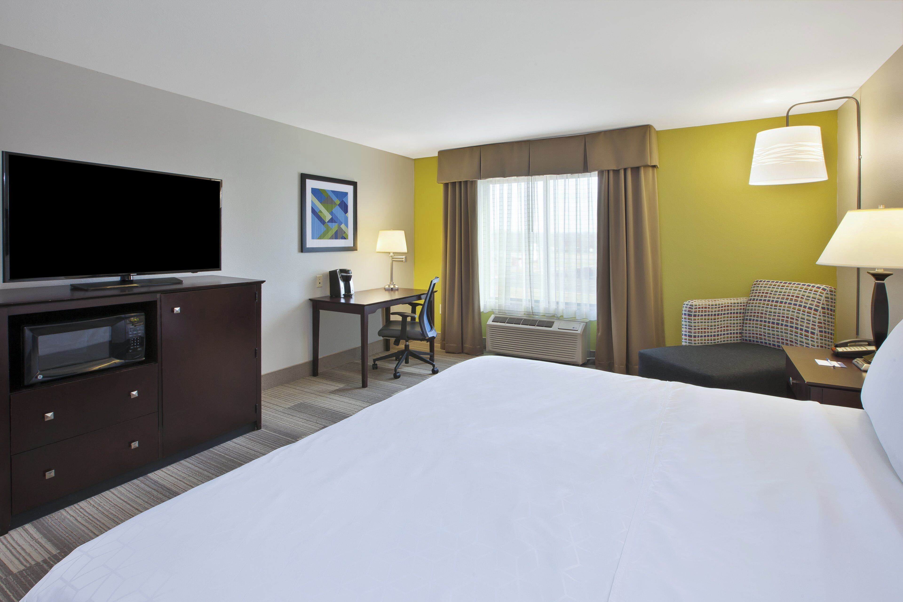 A hotel room with a large bed, a desk with a chair, a television, a microwave, a lamp, and an air conditioning unit.