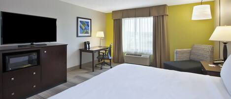 A hotel room with a large bed, a desk with a chair, a television, a microwave, a lamp, and an air conditioning unit.