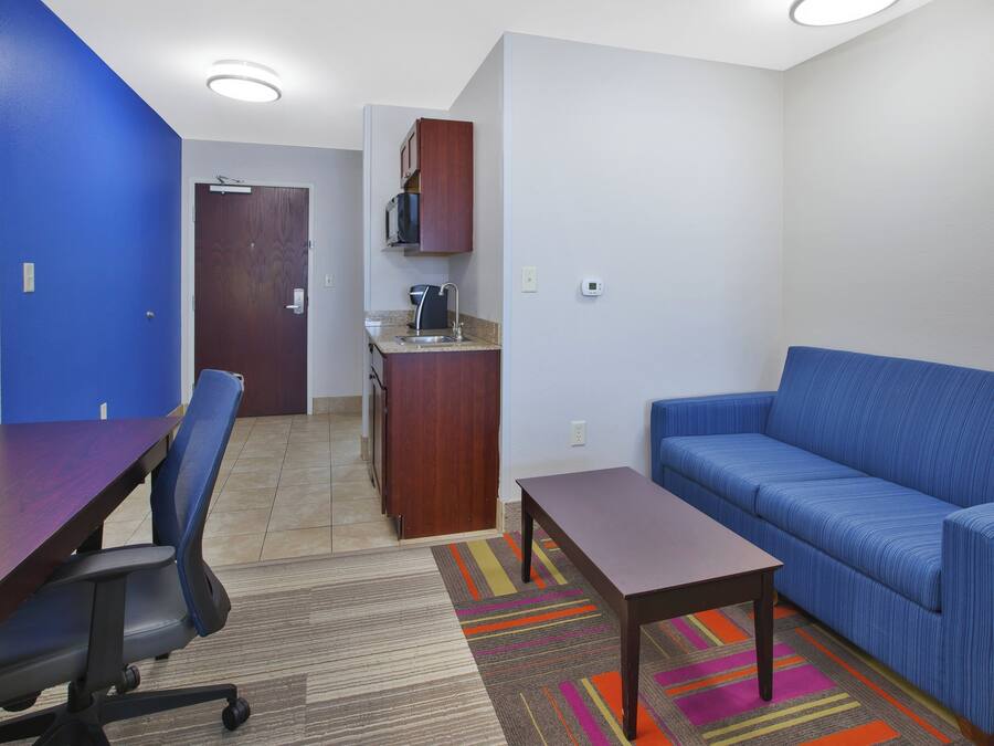A room with a blue sofa, a wooden table, a chair, a kitchenette, and a door.