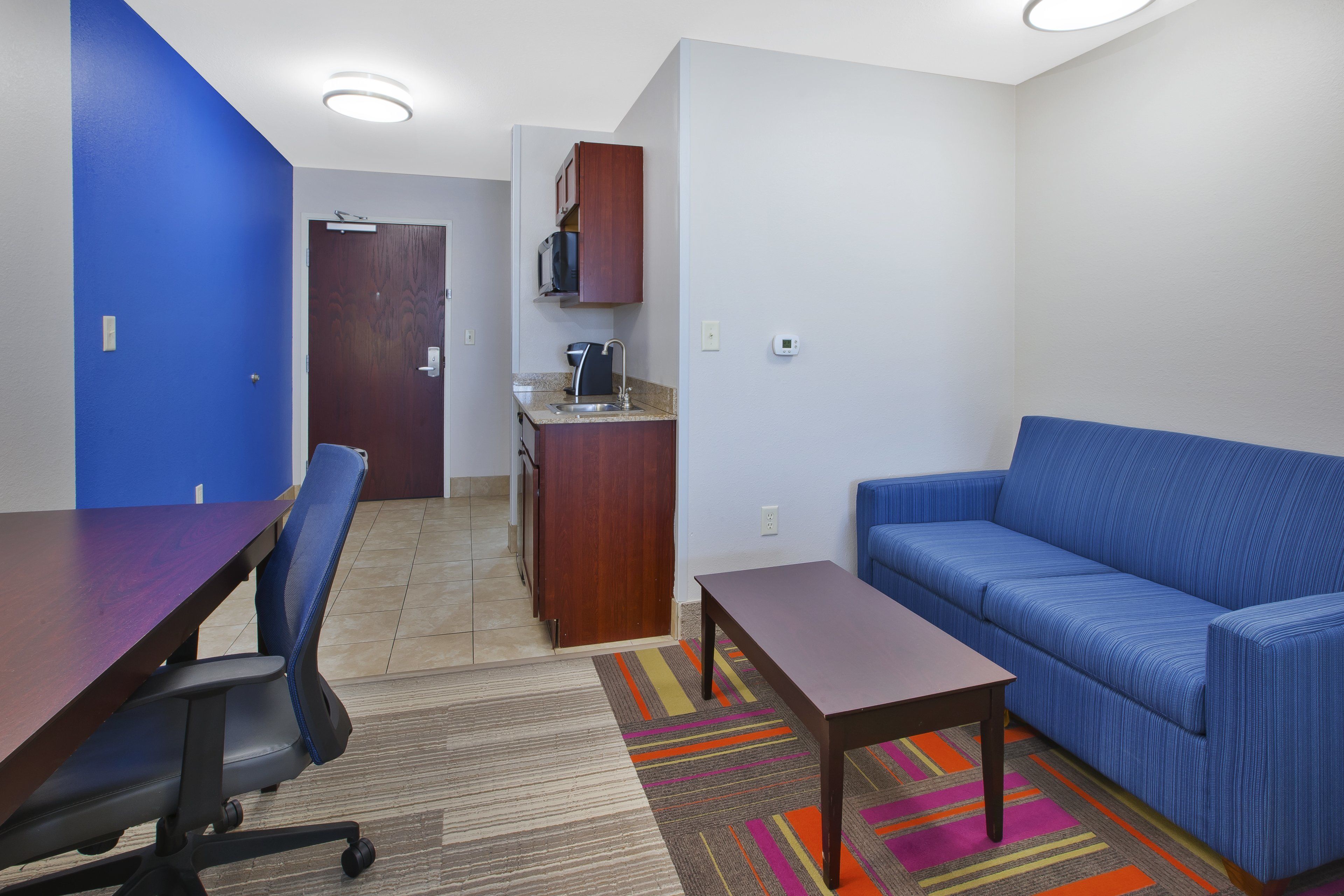 A room with a blue sofa, a wooden table, a kitchen area with a microwave, and a door leading to another room.