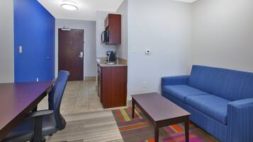 A room with a blue sofa, a wooden table, a kitchen area with a microwave, and a door leading to another room.