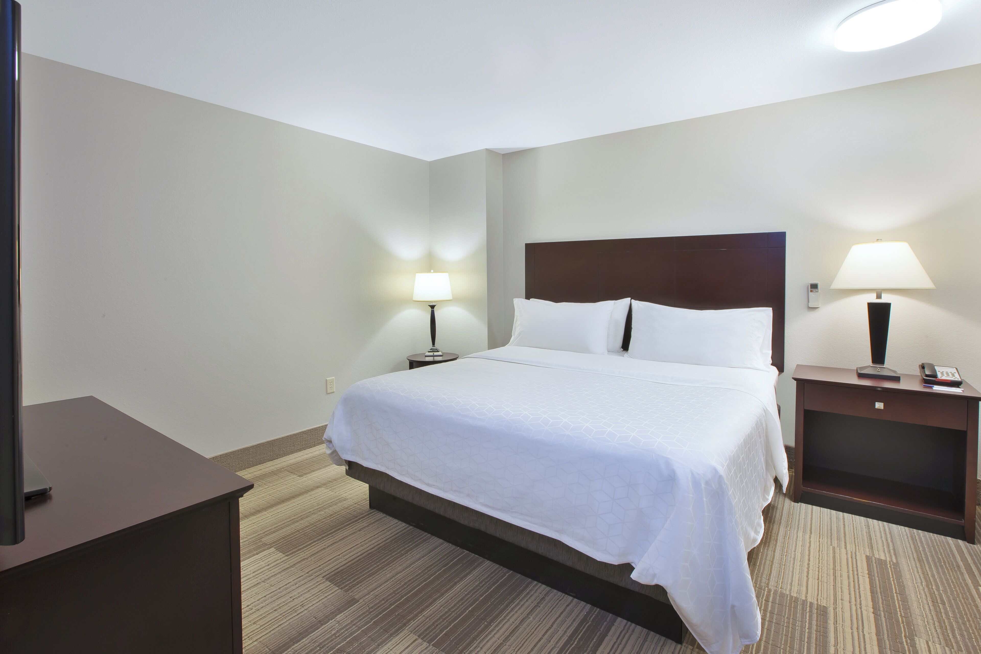 A hotel room with a large bed, two bedside tables with lamps, a TV on a stand, and a carpeted floor.