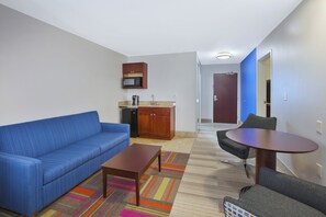 Een knus woongedeelte met een blauwe bank, een klein houten tafeltje, een kitchenette met een magnetron en een deur naar een andere kamer.