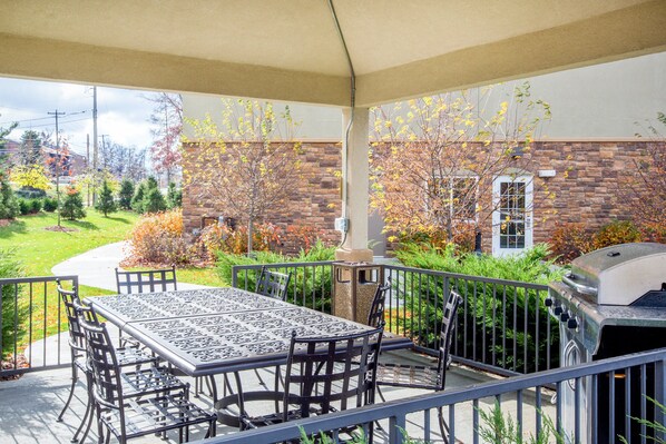 Een terras buiten met een tafel, stoelen, een barbecue en uitzicht op een bakstenen gebouw en tuin.