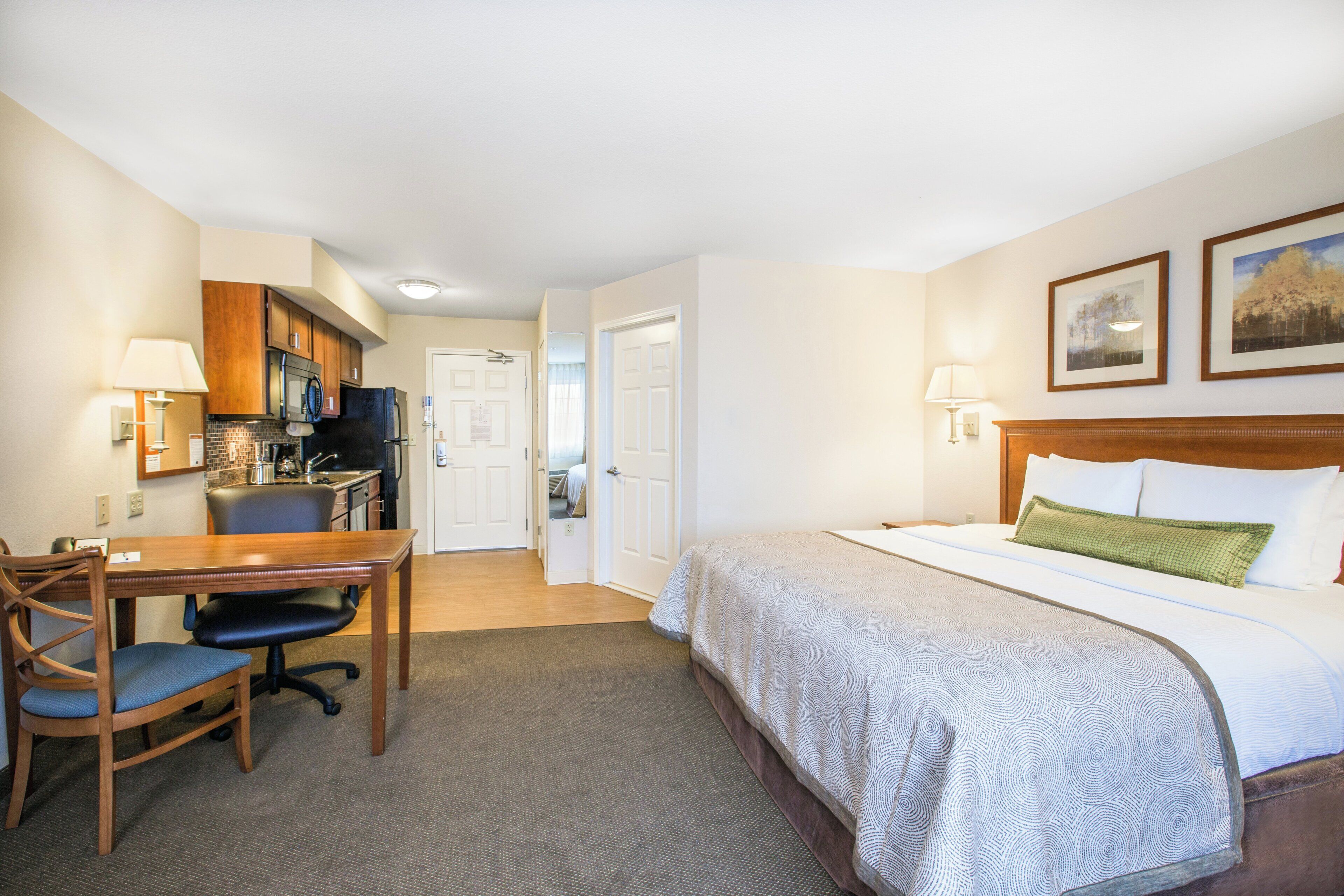 A hotel room with a bed, desk, chair, kitchenette, and a door leading to another room.