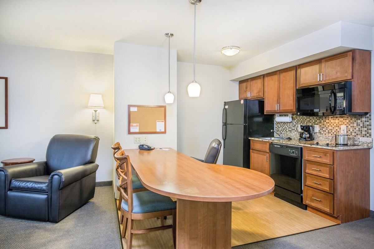 Cocina compacta con zona de comedor, que incluye una mesa y sillas de madera, refrigerador, microondas y cafetera.