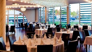 Ein Restaurant mit runden Tischen, Stühlen, Kronleuchtern und großen Fenstern.