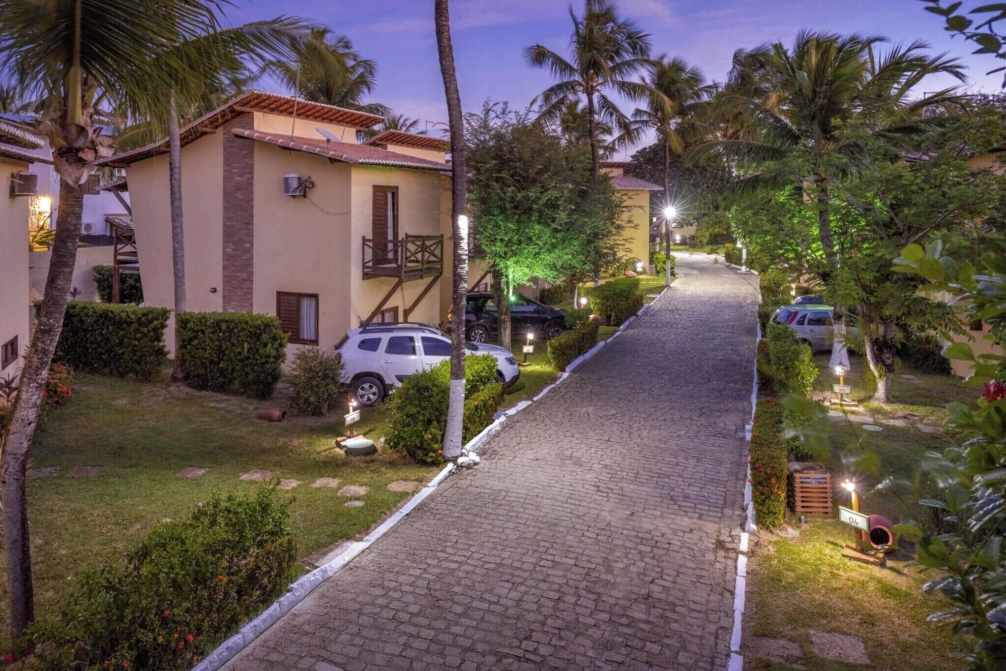 A paved pathway leading to a building with a balcony, surrounded by palm trees and greenery.