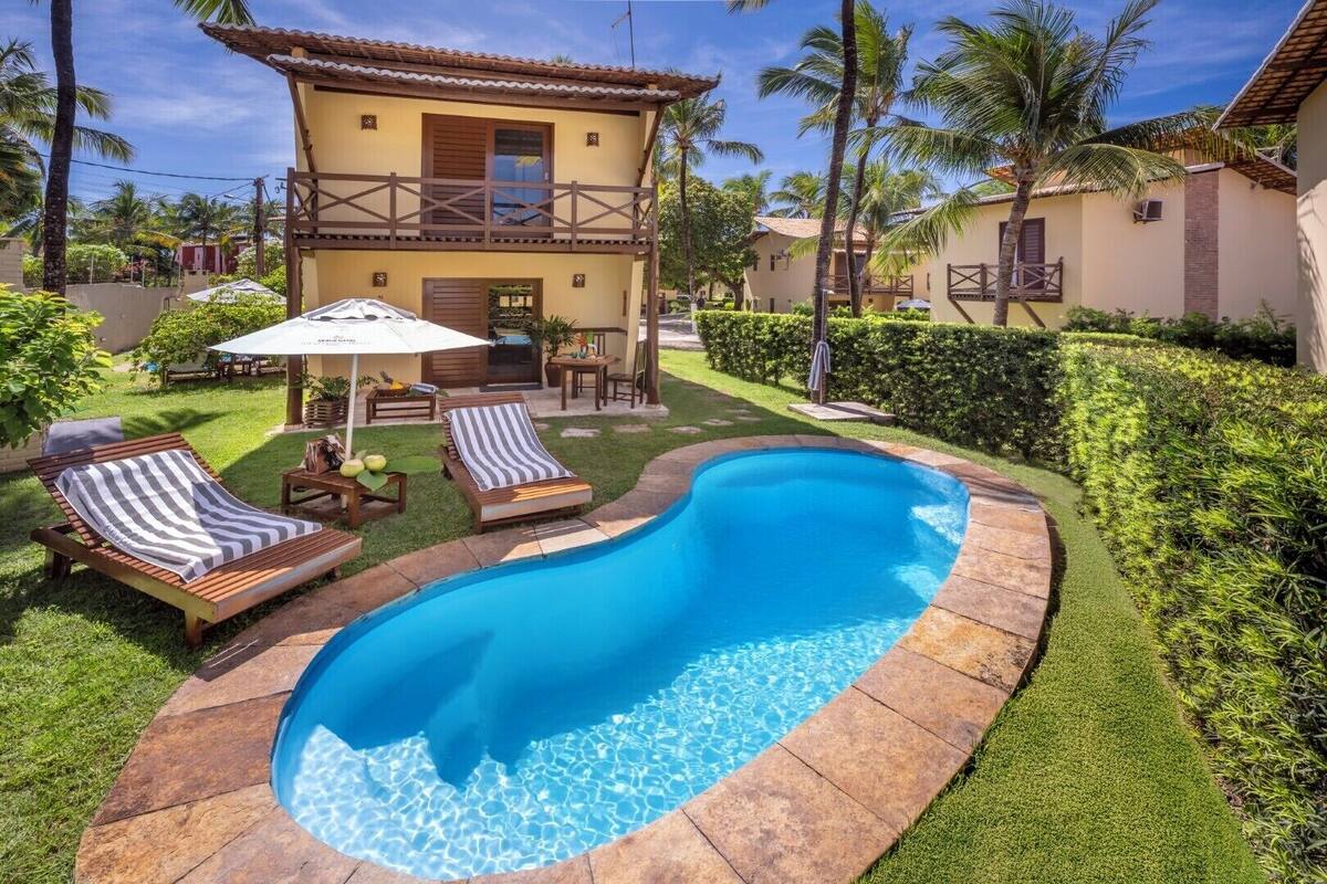 A swimming pool surrounded by lounge chairs and a patio area with a view of a yellow building and palm trees.