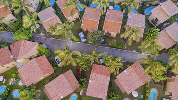 An aerial view of a resort with multiple bungalow-style buildings, each with a tiled roof and surrounded by palm trees. There are several swimming pools visible, indicating a resort setting.