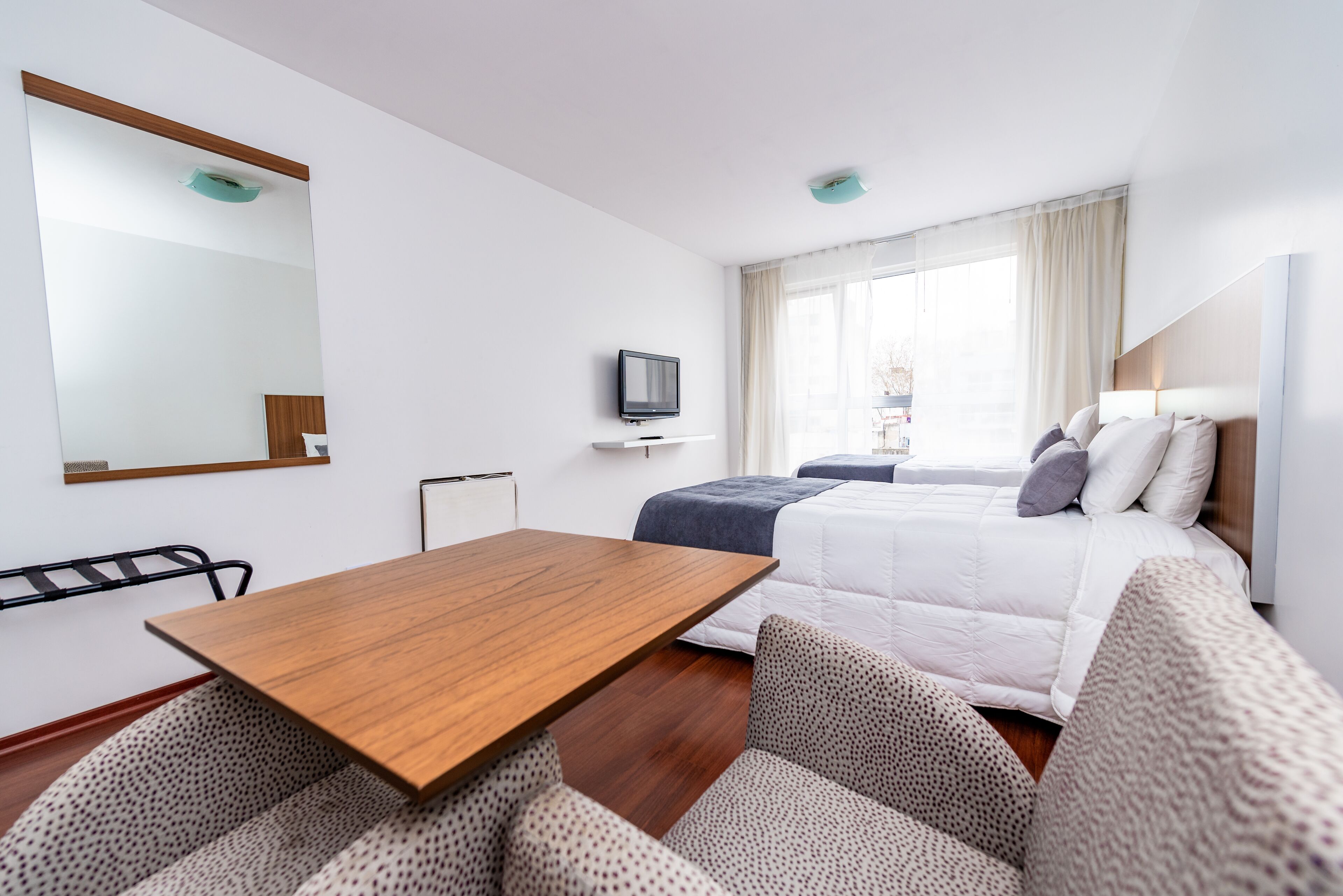 A hotel room with a wooden table, a bed with a white duvet, a TV on a stand, and a large window with curtains.