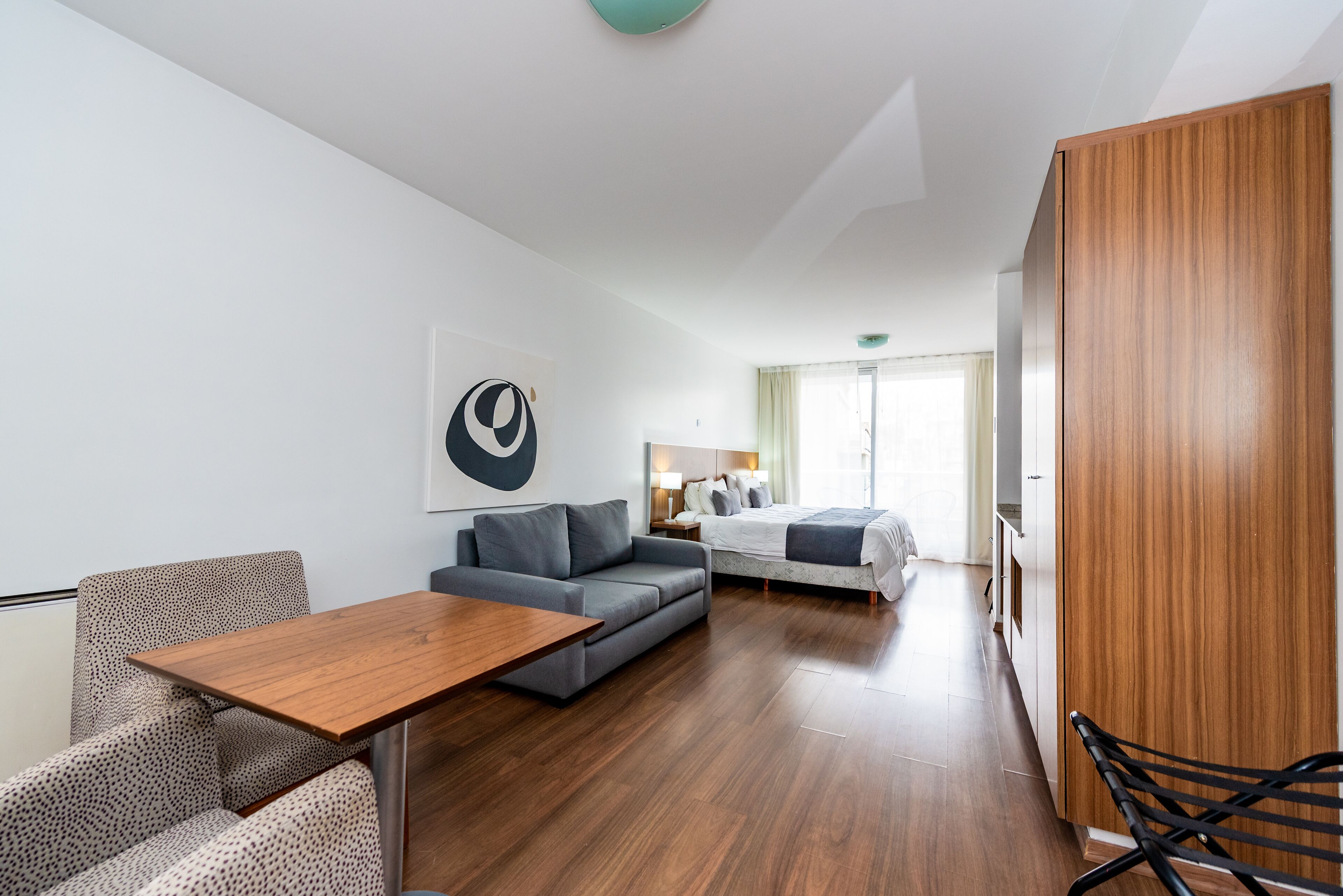 Ein modernes Hotelzimmer mit Holzboden, einem grauen Sofa, einem kleinen Tisch, einem Bett mit grauer Daunendecke und einem Holzschrank.
