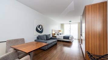 Ein modernes Hotelzimmer mit Holzboden, einem grauen Sofa, einem kleinen Tisch, einem Bett mit grauer Daunendecke und einem Holzschrank.