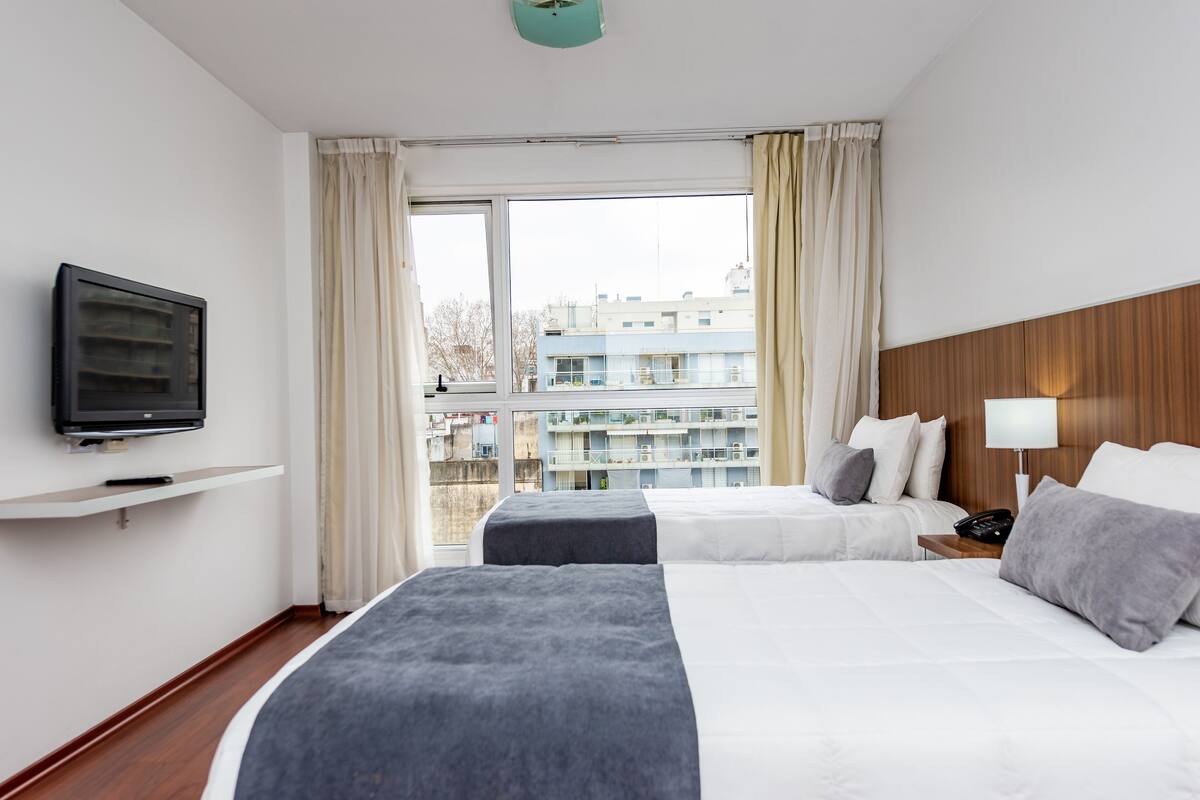 A hotel room with two beds, a TV on a shelf, a bedside table with a lamp, and a window with curtains.