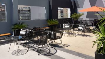 Um pátio externo com cadeiras e mesas de metal preto, plantas em vasos e um grande guarda-sol laranja.