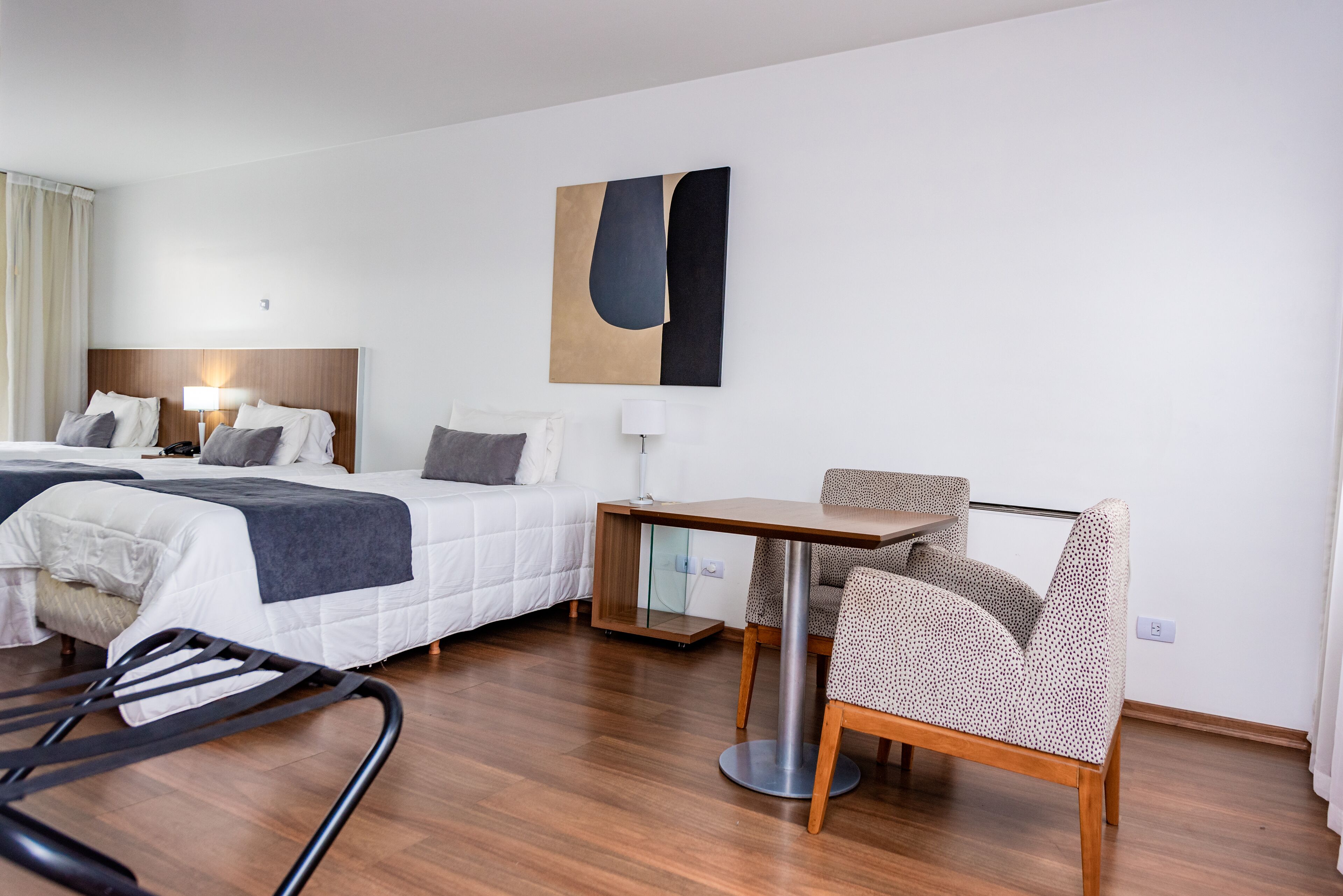 A hotel room with two beds, a small table, a chair, and a large abstract painting on the wall.