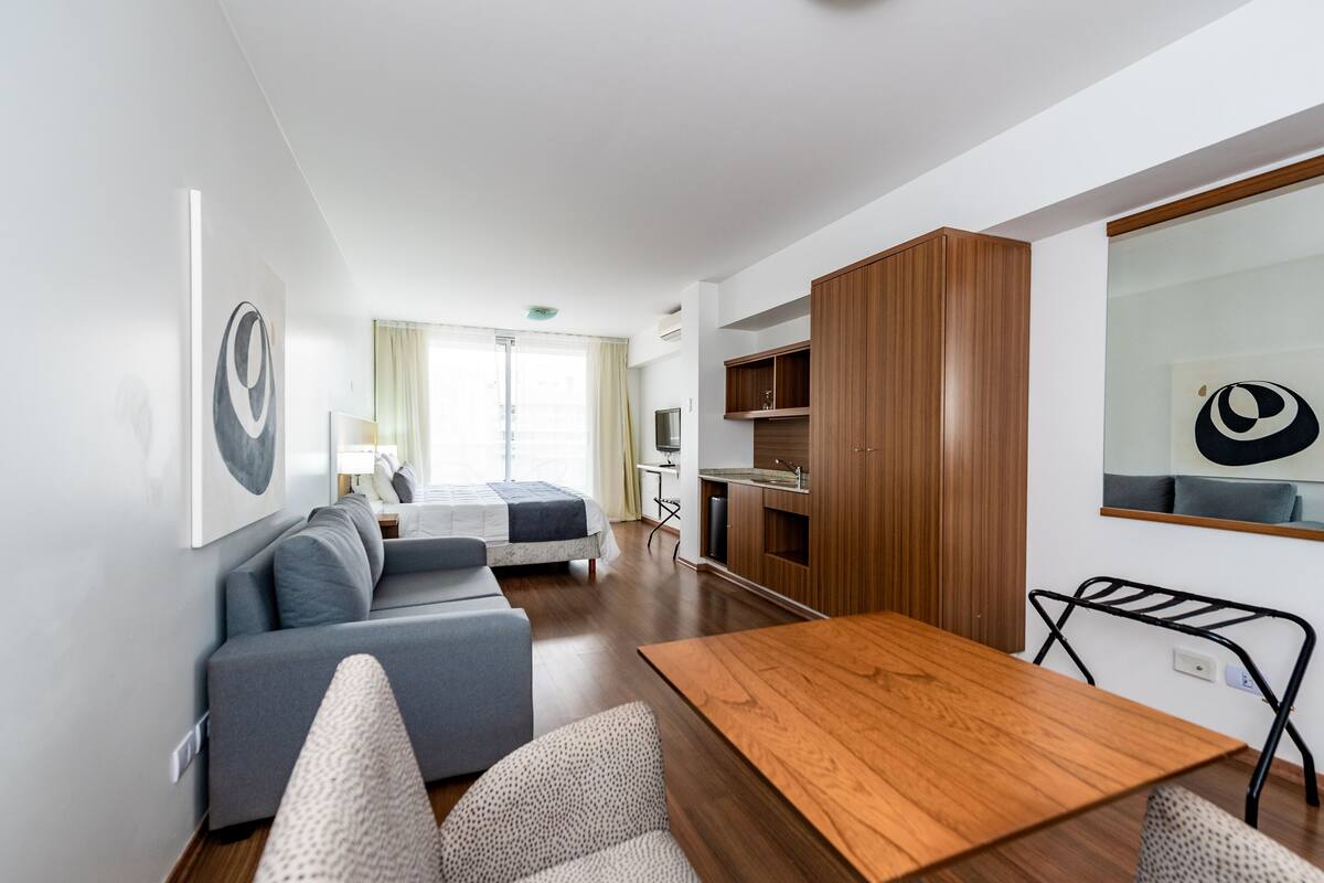 A modern, compact living space with a wooden dining table, grey sofa, and a kitchenette area.