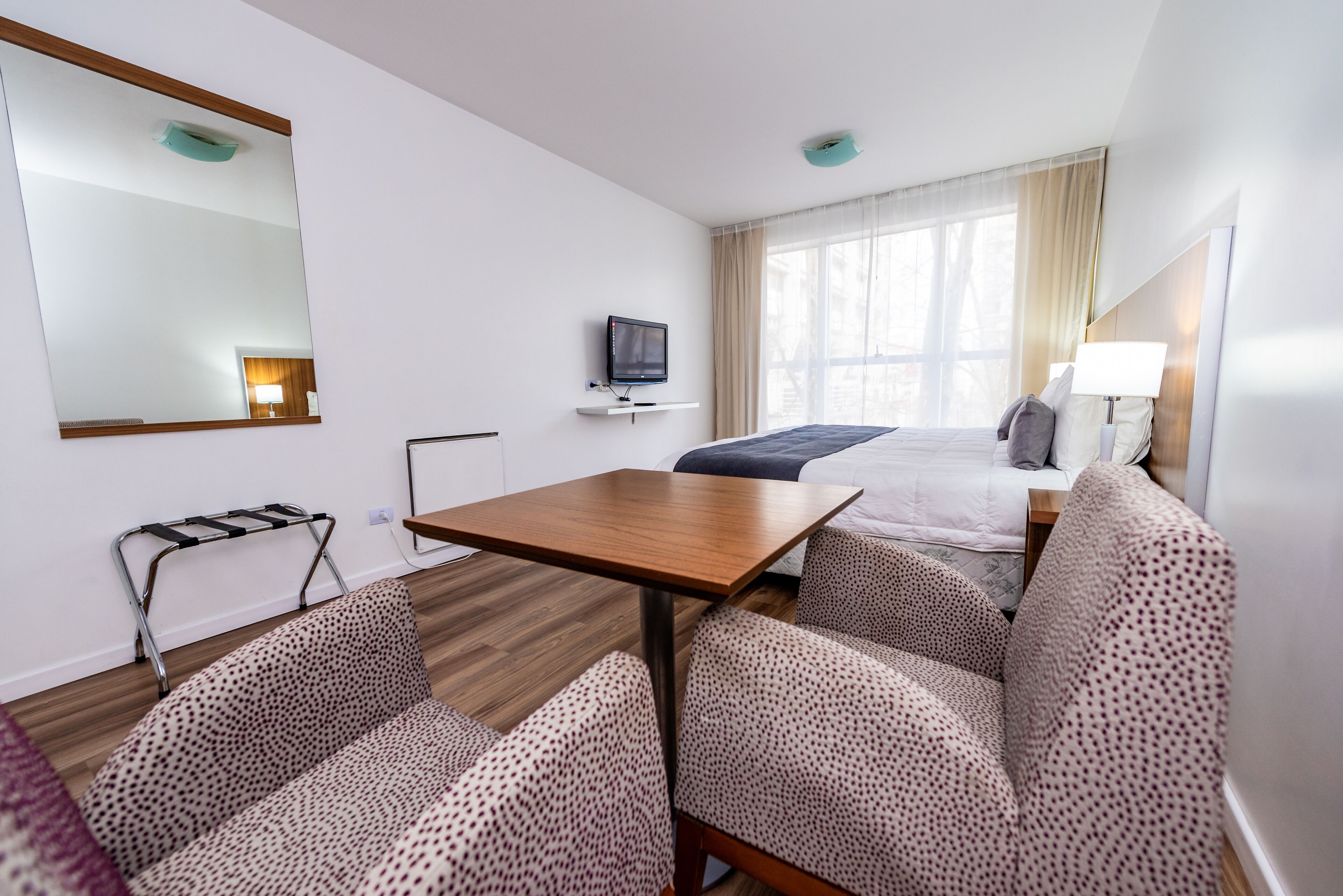 A hotel room with a wooden table, two patterned sofas, a bed with a blue bedspread, a TV on a shelf, and a large mirror.